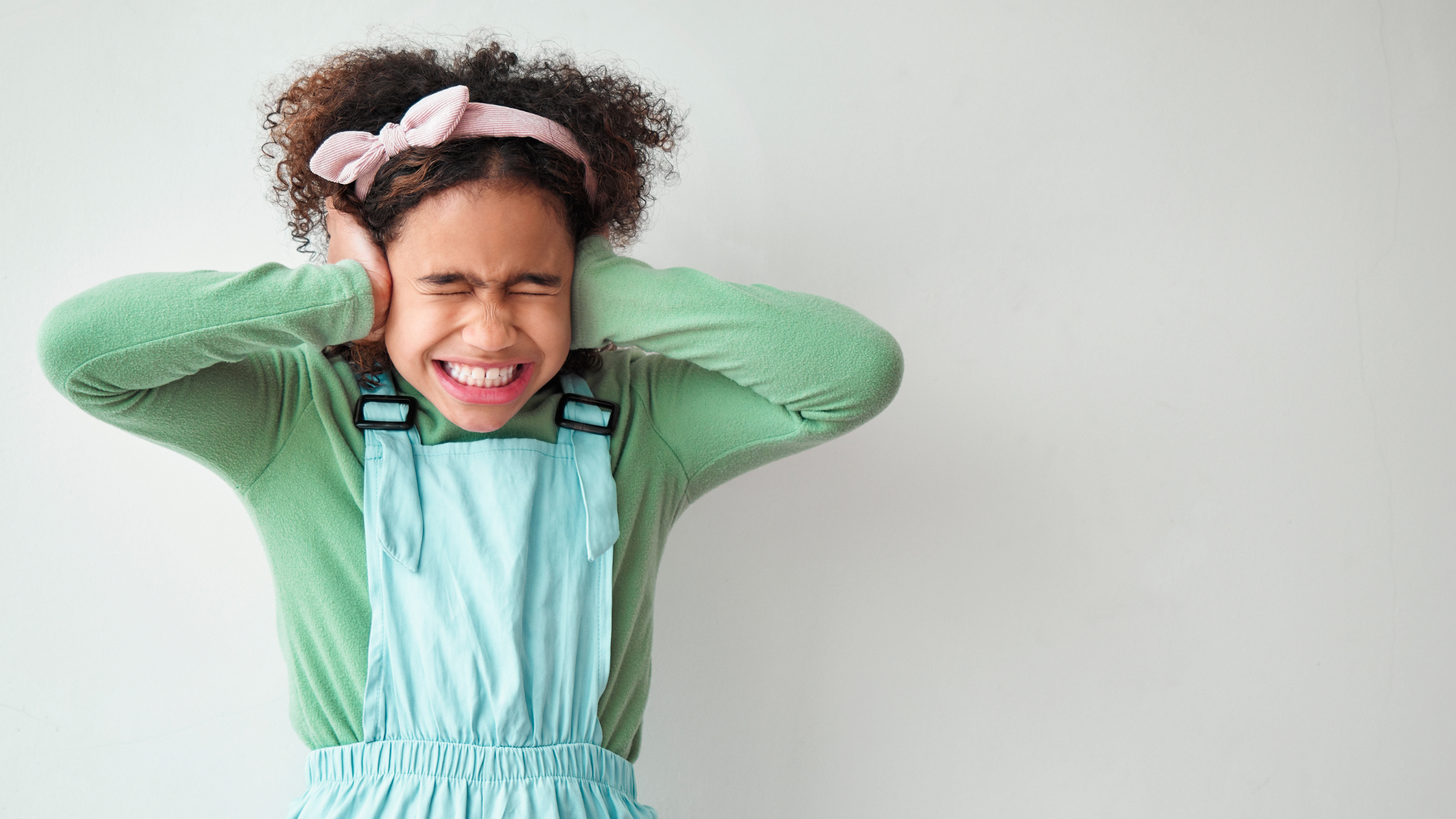 Young girl wearing a green jumper and dungarees, holding her ears